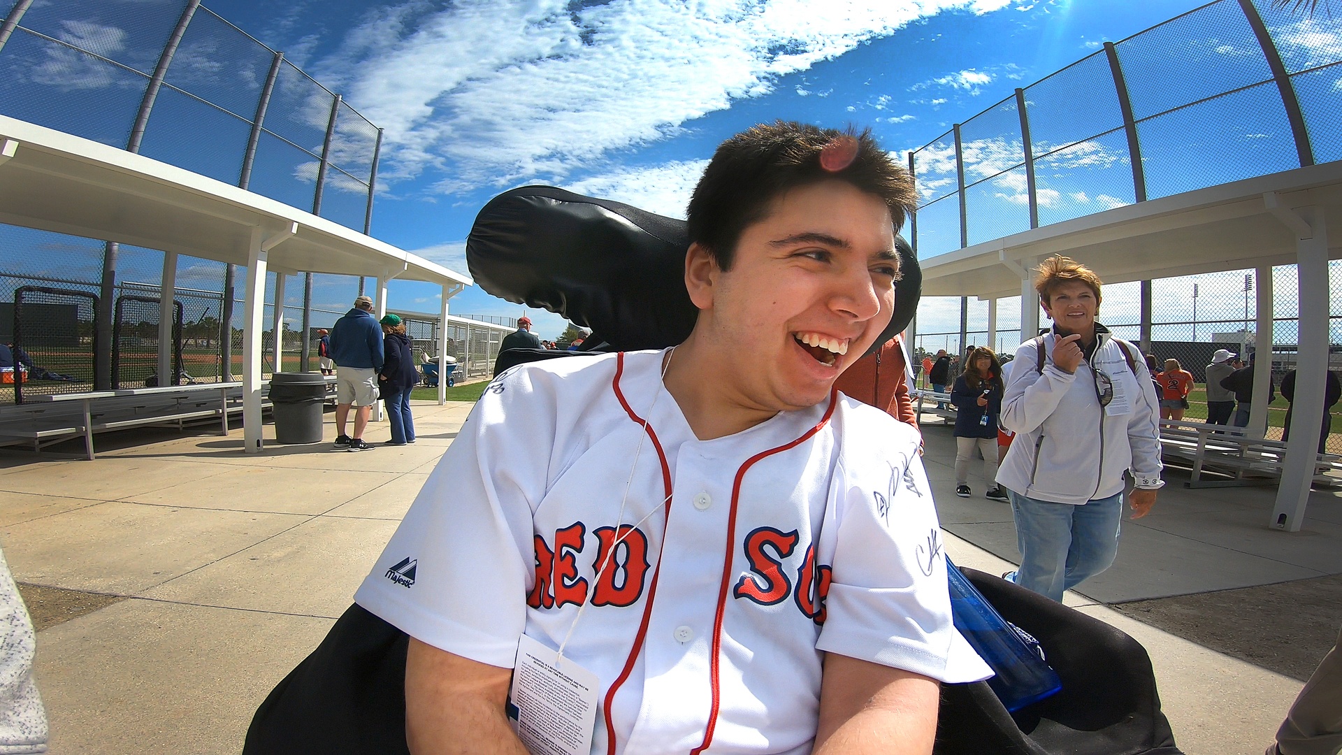 The image shows a man wearing a white Red Sox baseball jersey who appears to be in a motorized wheelchair. He has short brown hair and is smiling and is possibly engaged in an outdoor athletic game, most likely baseball.